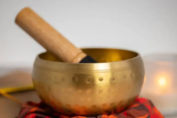 A front and close-up view of a brass metal Tibetian singing bowl with a lit candle is seen blurry in the background.