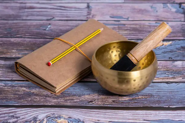 A close-up view of a brass metal Tibetian singing bowl on a blue wooden table with a notebook and a pencil next to it.