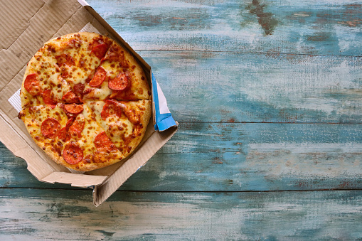 Stock photo showing elevated view of cardboard takeaway pizza box containing a sliced pepperoni pizza, topped with a rich tomato sauce and melted buffalo mozzarella on blue wood grain background.