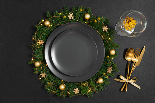 Christmas table setting with empty black plate, golden cutlery and a glass on black background. New Year party. Copy space, top view, flat lay.