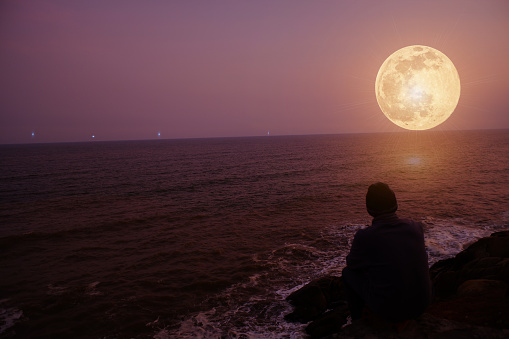 Man in silhouette looking at the distance at the full moon and silver stars overseas with a calm orange sky.