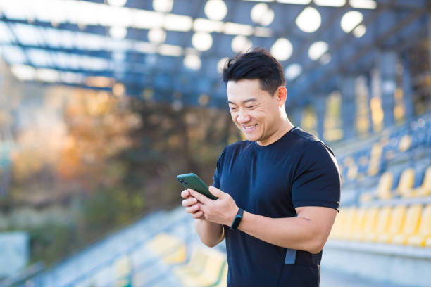 A healthy man watches a video on a mobile phone after a workout while sitting outdoors using an app on a 4g wireless device. Asian athlete is happy and smiling after running rest A healthy man watches a video on a mobile phone after a workout while sitting outdoors using an app on a 4g wireless device. Asian athlete is happy and smiling after running rest scoring run stock pictures, royalty-free photos & images