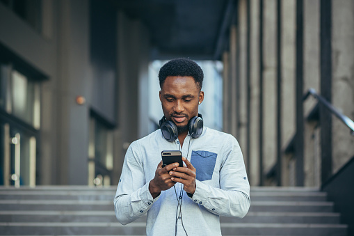 Young afro american student man listens to podcast using app on phone, and big earphone, in casual clothes of the day