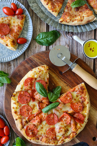 Stock photo showing elevated view of circular, wooden chopping board containing a sliced pepperoni  pizza and a paper plate of Pizza Margherita, both topped with a rich tomato sauce, melted buffalo mozzarella and garnished with fresh basil leaves.