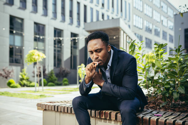 african american business man smoking cannabis outdoors sitting on city park bench on urban street background. Male employee Businessman. Office worker in suit Relieves stress a marijuana outside african american business man smoking cannabis outdoors sitting on city park bench on urban street background. Male employee Businessman. Office worker in suit Relieves stress a marijuana outside relieves stock pictures, royalty-free photos & images