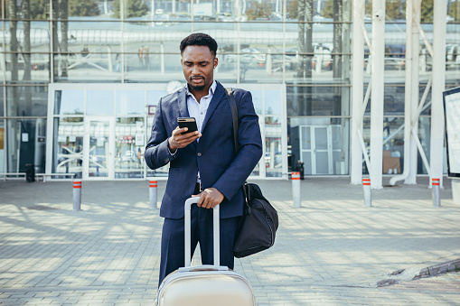 african businessman traveling near airport with suitcase calling taxi using mobile app