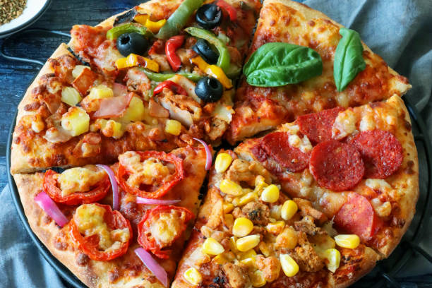 Stock photo showing close-up, elevated view of triangular slices of pizzas with different toppings including Hawaiian (ham and pineapple), feta cheese, tomato and red onion, chicken and sweetcorn, pepperoni, vegetable (yellow and green bell pepper with black olives) and Pizza Margherita (melted golden buffalo mozzarella cheese, rich tomato marinara sauce and fresh basil leaves).