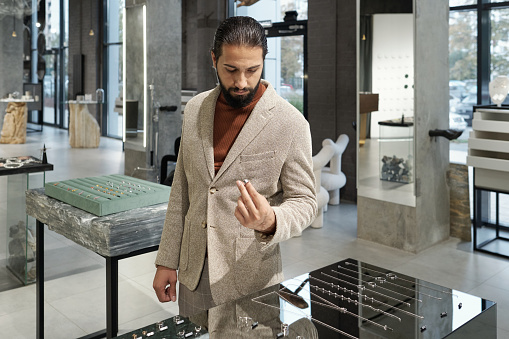 Young serious bearded man in formalwear choosing ring in jewelry shop while standing by display with large assortment