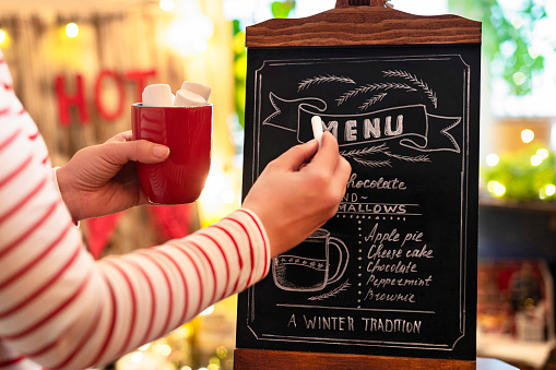 Woman with cup of cocoa with marshmallows drawing christmas menu of cocoa bar on small chalk board.
