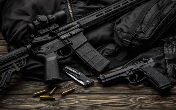istock Modern automatic rifle with a telescopic sight on a dark background. Jacket, cap, gloves on a wooden table. The uniform of a guard or a mercenary. 1358418296