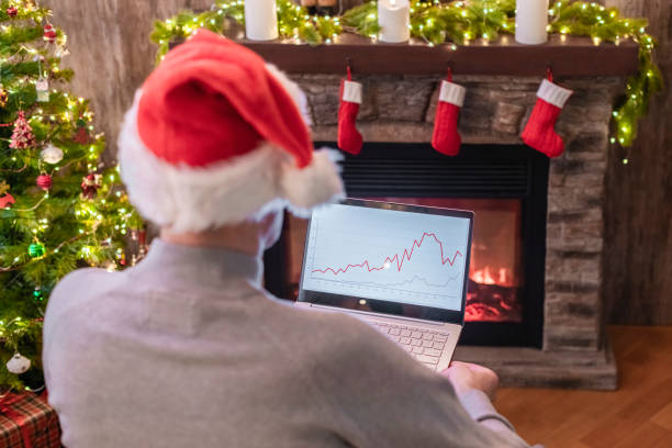 istock Man freelancer in santa claus hat working on laptop with graphs and charts on screen sitting near christmas tree and fireplace. 1358419111