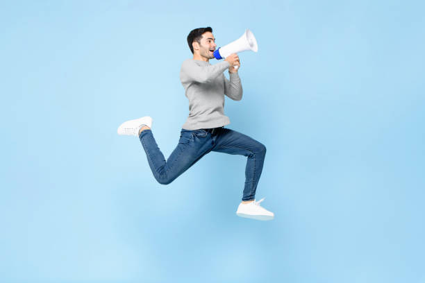 Portrait of energetic young Caucasian man jumping with megaphone isolated on light blue background stock photo