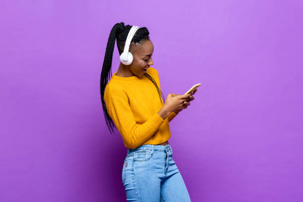 African American woman wearing headphones and listening to music from smart phone on colorful purple isolated studio background stock photo