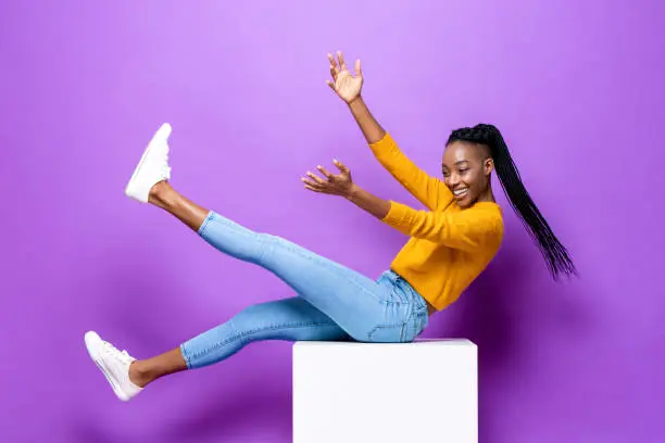 Smiling young African-American woman lying on stool raising hands and leg in studio purple color isolated background
