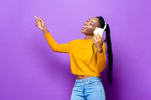 African American woman dancing and singing while listening to music from smart phone on colorful purple isolated studio background