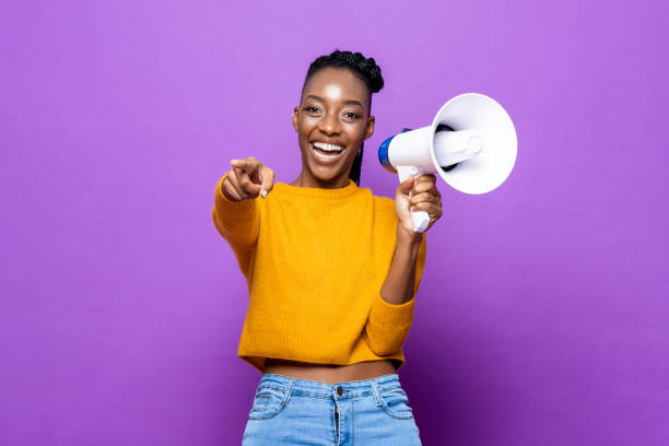 mujer afroamericana sonriente sosteniendo megáfono y señalando con el dedo en un fondo de estudio púrpura aislado - contar fotografías e imágenes de stock