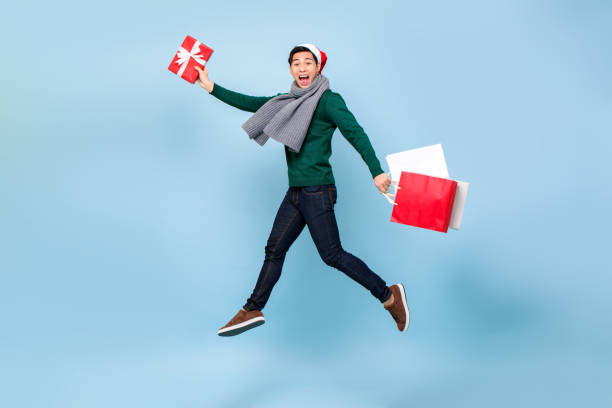 retrato completo de um jovem asiático saltitante com sacolas de compras e caixa de presente de natal em fundo isolado de estúdio azul claro - studio sho - fotografias e filmes do acervo