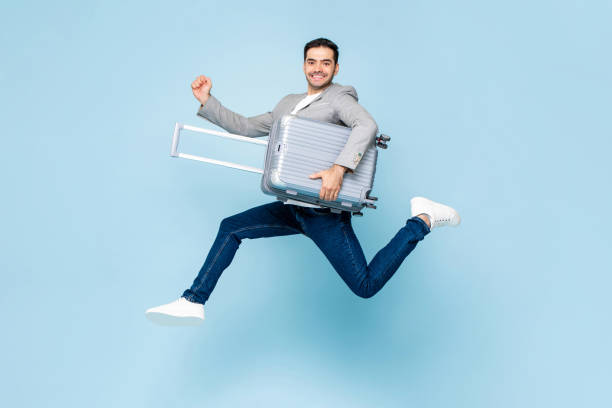 Young happy handsome Caucasian tourist man with baggage jumping in isolated light blue studio background