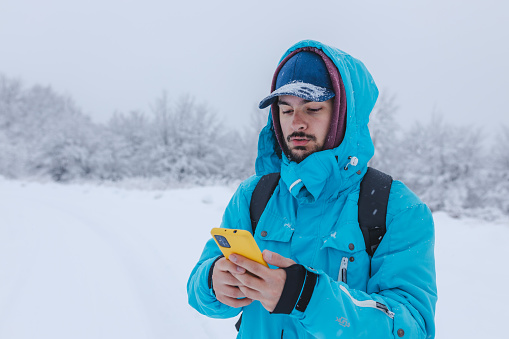 Young male hiker using his smartphone while being on top of a mountain during snowfall