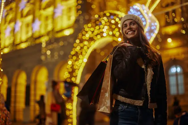 Smiling young adult woman walking through the city streets at Christmas, going from shopping and carrying shopping bags with Christmas gifts