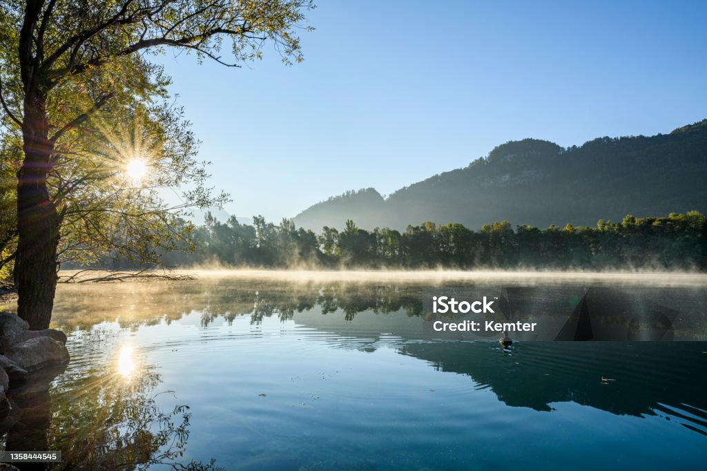 Sunrise at a idyllic lake Sunrise at a idyllic lake with calm water and some fog above it and the reflection of the mountains. A loon is swimming on the water. Vorarlberg, Nuziders Austria Stock Photo