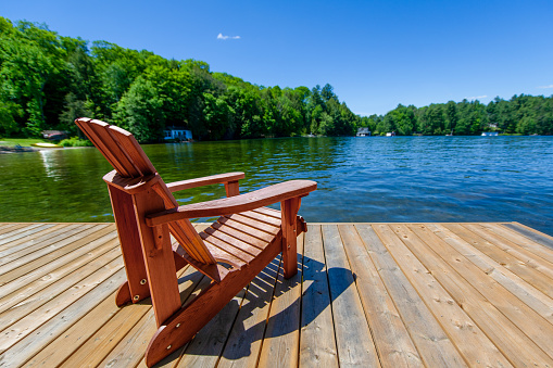 Brown Adirondack chair on a wooden pier facing a lake in Muskoka, Ontario Canada. Across the calm water are few cottages nestled among green trees.