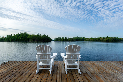 Two white Adirondack chairs on a wooden dock facing the blue water of a lake in Ontario Canada. Cottages nestled among green trees are visible across the water.