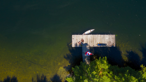 Aerial view of a cottage wooden pier on a lake in Muskoka, Ontario Canada. Two brown Adirondack chairs are visible on the dock facing the blue waters of the lake.
