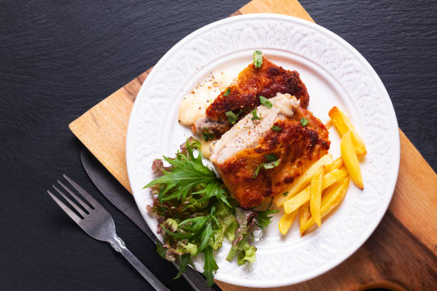 speisekonzept hausgemachtes kalbskordon bleu mit salat und französisch gebraten in weißem keramikteller auf holzbrett und schwarzem schiefersteinhintergrund mit kopierraum - cordoning stock-fotos und bilder