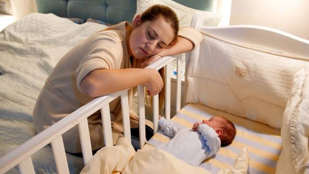 la joven madre cansada y agotada se quedó dormida mientras mecía la cuna de su bebé recién nacido por la noche. concepto de noches de insomnio y depresión de los padres después del parto - cot fotografías e imágenes de stock