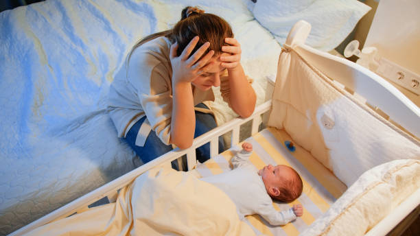 une jeune mère fatiguée souffrant de dépression regarde son nouveau-né insomniaque dans un berceau la nuit. dépression maternelle après l’accouchement et nuits blanches. - domestic life sadness family bed photos et images de collection