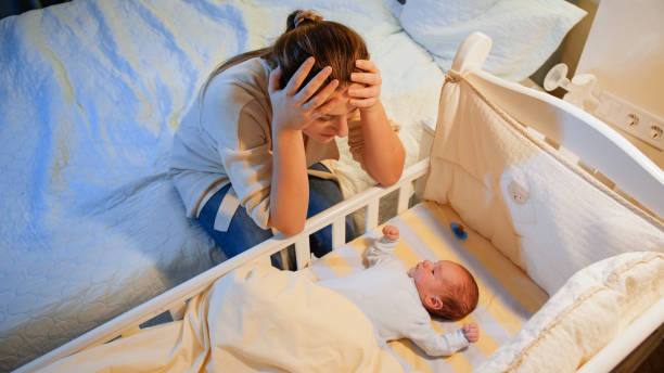 Young tired mother suffering from depression lookin on her sleepless newborn baby in crib at night. Maternal depression after childbirth and sleepless nights. Young tired mother suffering from depression lookin on her sleepless newborn baby in crib at night. Maternal depression after childbirth and sleepless nights postpartum depression stock pictures, royalty-free photos & images