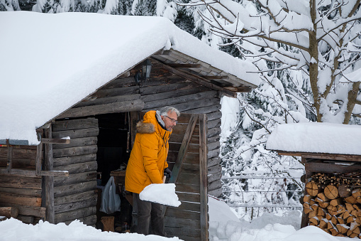 Elder man shovels snow in front of the cottage. Preparing for Christmas decoration.