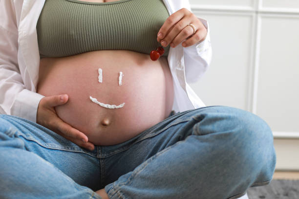 Close-up of torso of young pregnant model applying moisturizer on her belly to avoid stretch marks. Future mom with funny smile from moisturizing cream on her tummy. Pregnancy skincare concept. primer plano del torso de la joven modelo embarazada aplicándose crema hidratante en el vientre para evitar las estrías. futura mamá con sonrisa divertida de crema hidratante en su barriga. concepto de cuidado de la piel durante el embarazo