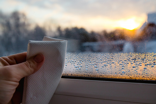 Vista de cerca de la mano de la persona usando tela de papel, secando gotas de condensación húmedas de la ventana de vidrio en la fría mañana de invierno al amanecer. photo