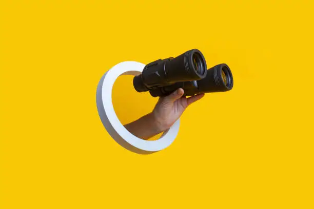 Photo of Woman's hand holding binoculars in a hole on a yellow background.