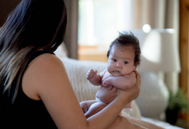 An indigenous mother sits her newborn daughter on her lap as she holds her out to get a good look at her features.  She is dressed casually and the two are seated on the bed as the mother looks over the baby and talks gently to her.