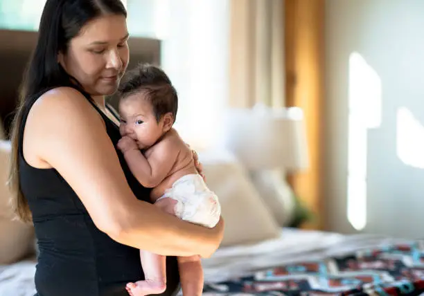 An indigenous mother holds he newborn daughter closely to her chest as they enjoy some bonding time.  The mother is dressed casually in a black tanks top and is seated on her bed ad=s she enjoys some close skin-on-skin time with the diapered infant.