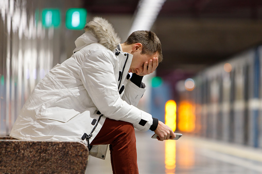 Crying man in white parka very upset, holding smartphone, gets bad news, covers his face with his hand, sitting on bench in subway station. Problem in relationships, love depression, loneliness