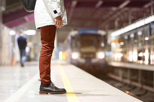 Close up of man waiting train at metro station early morning and goes to work, dressed in white parka, red jeans, brown leather boots, empty subway platform on background, bottom view.