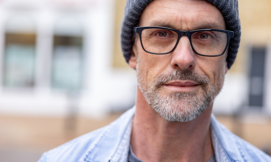 Close-up portrait of an adult man on the street wearing glasses and a beanie and looking at the camera