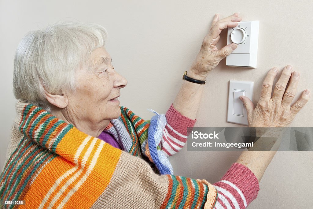 Senior woman adjusting her thermostat Senior woman saving energy by dressing warm and adjusting her thermostat. Senior Adult Stock Photo
