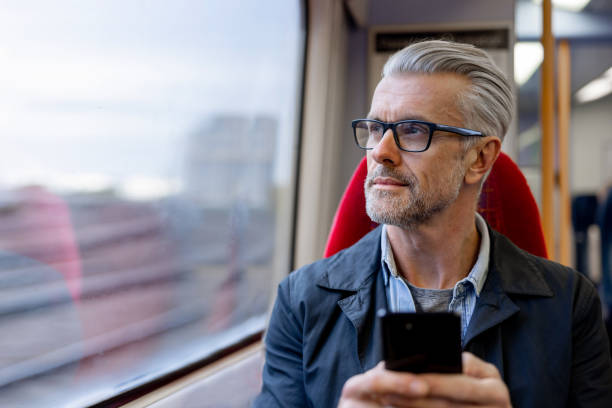 uomo premuroso che usa il suo telefono mentre viaggia su un treno - white hair foto e immagini stock