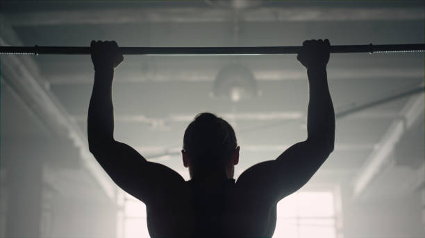 Man doing wide grip pull ups on bar. Athlete pumping arm muscles with chin ups Back silhouette muscular man pull ups exercise on horizontal bar. Closeup powerful athlete pumping arm muscles at pullups workout in dark gym. Strong bodybuilder training biceps muscles strength gripping bars stock pictures, royalty-free photos & images