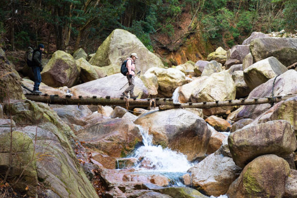Two Japanese hikers on their way up a mountain trail cross a small wooden bridge over a waterfall stream.