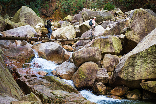 Two Japanese hikers on their way up a mountain trail cross a small wooden bridge over a waterfall stream.