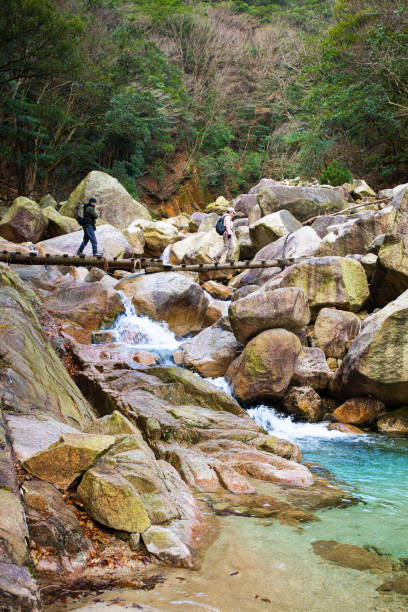 Two Japanese hikers on their way up a mountain trail cross a small wooden bridge over a waterfall stream.