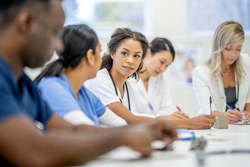 A small group of medical students sit together at a table as they individually study. Each one is dressed professionally in medical attire and has books open in front of them as they works to earn their medical degrees. A few students are talking amongst themselves as they work to help each other.
