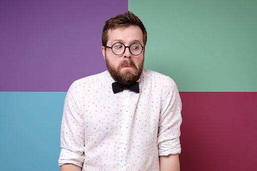 Bizarre, frightened man in round glasses, a shirt and a bow tie hunched shoulders in bewilderment and looks down with a strange grimace on face. Studio, multicolored background.