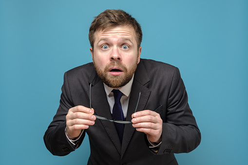 Shocked, frightened business man in a suit has taken off glasses and looks with big eyes opening mouth. Isolated on a blue background.